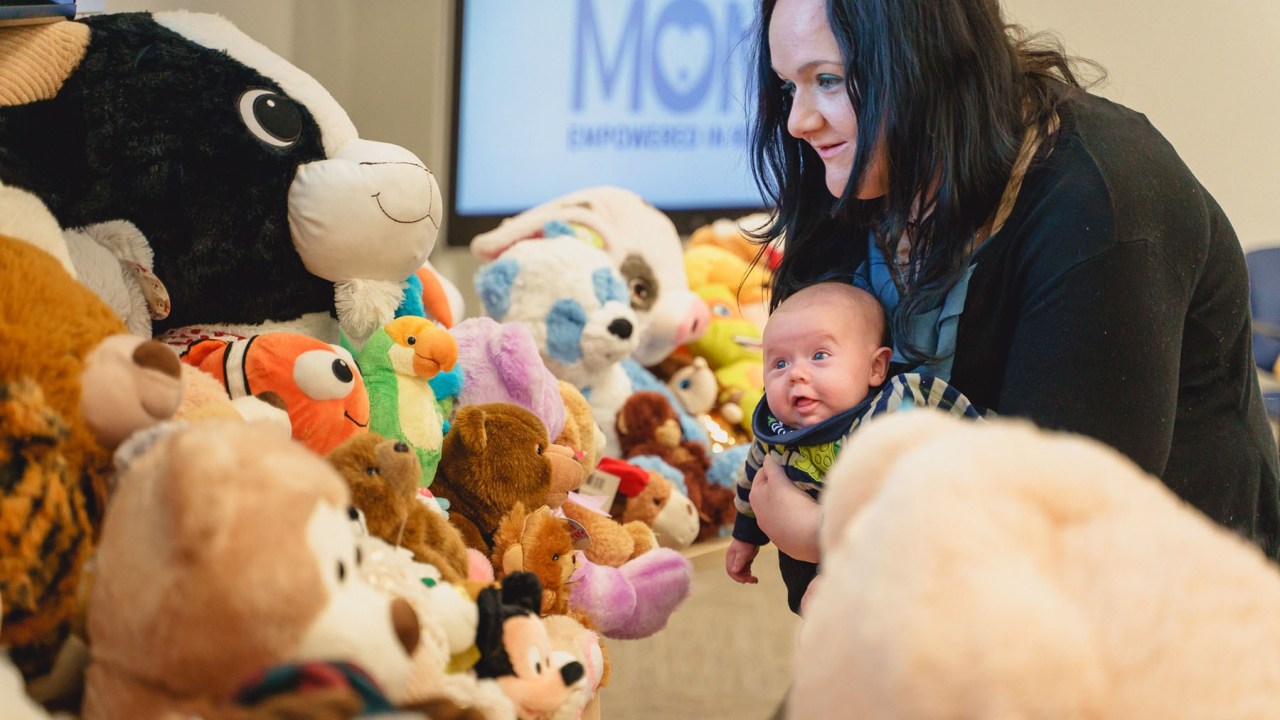 A mother holding a baby and looking at stuffed animals.