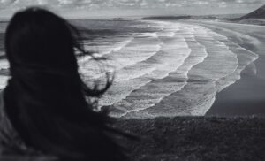 Black and white image of woman from back looking over waves breaking on the sand