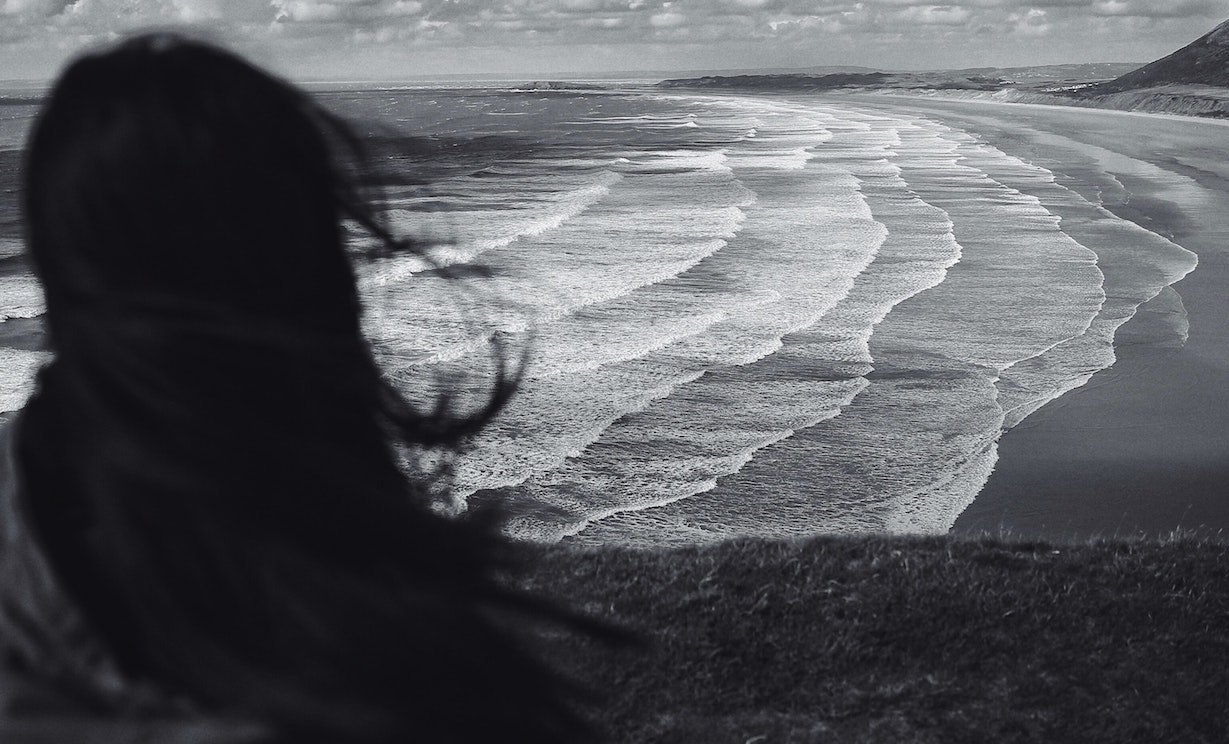 Black and white image of woman from back looking over waves breaking on the sand