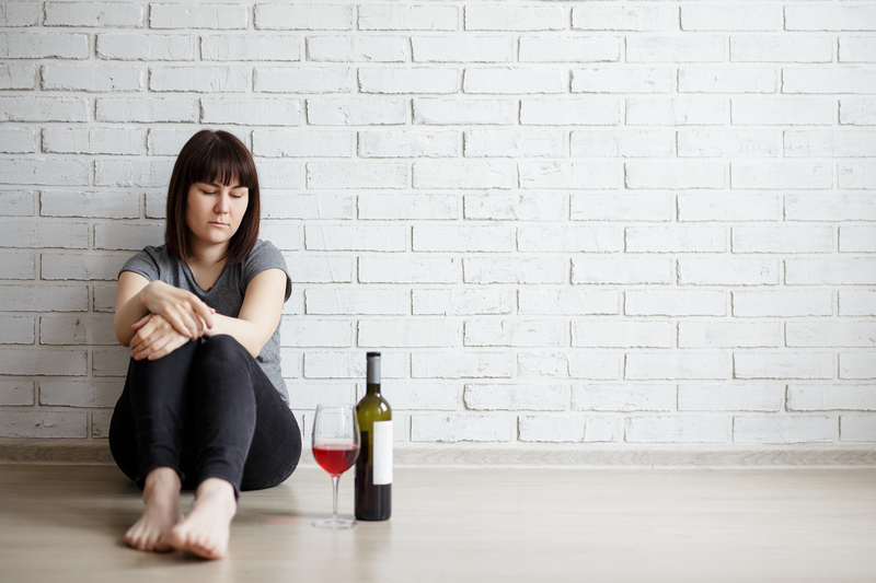 Woman sits on floor against wall, bottle and glass of wine next to her.