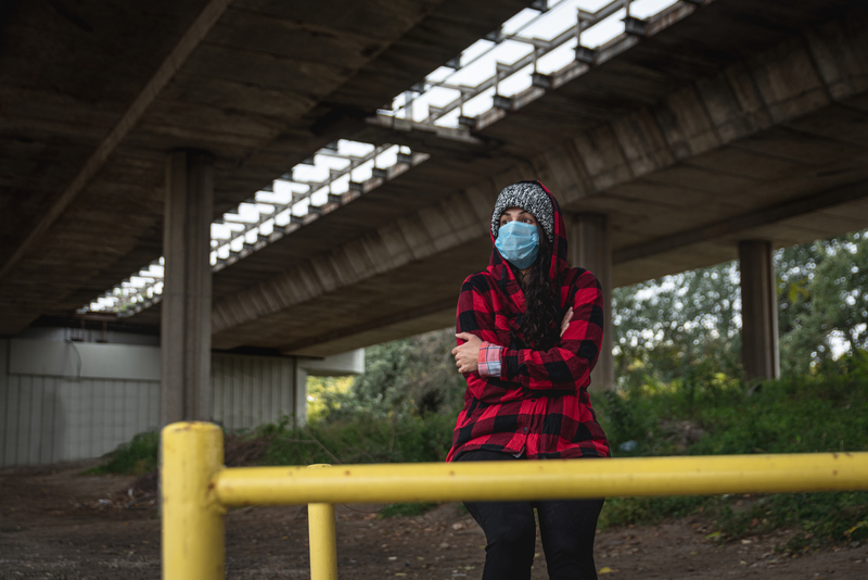 Woman wearing mask sitting alone under a bridge or freeway, bundled up.