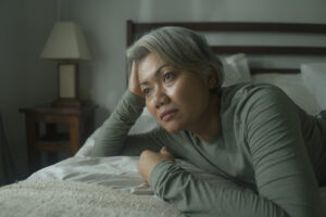 Woman with gray hair on bed, head resting on hand, sad.