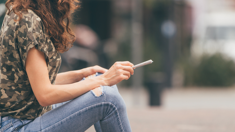 Person sitting on curb or ground and holding joint or rolled cigarette.