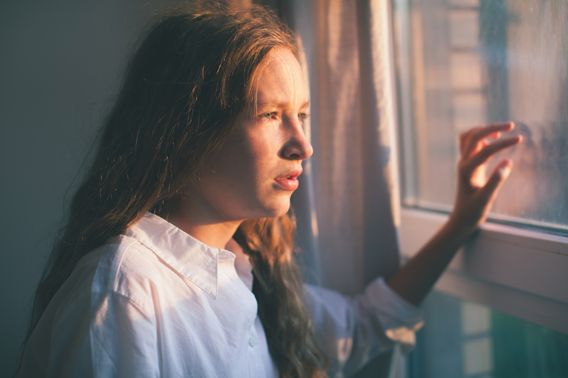 Girl staring out window with hand on window, sad.