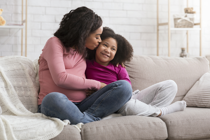 Mother kisses young daughter on the cheek. They are sitting cross-legged on a white couch.