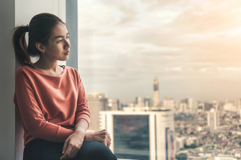 Woman sits on windowsill, looking out. She is serious or depressed.