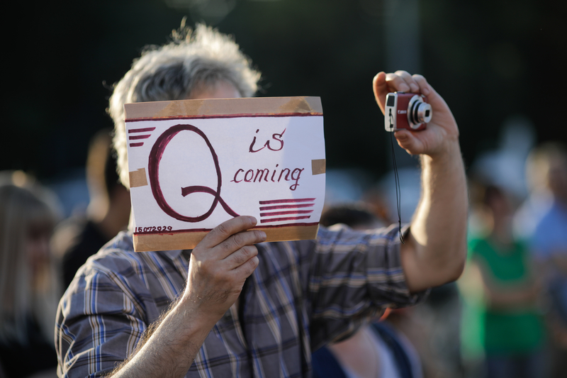 Man has sign covering his face that reads "Q is coming". He holds camera with other hand, at protest in Romania