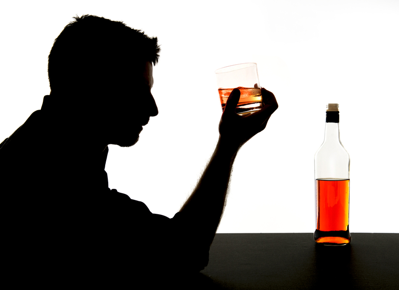 Silhouette of man contemplating drink in hand, with bottle of alcohol in front of him