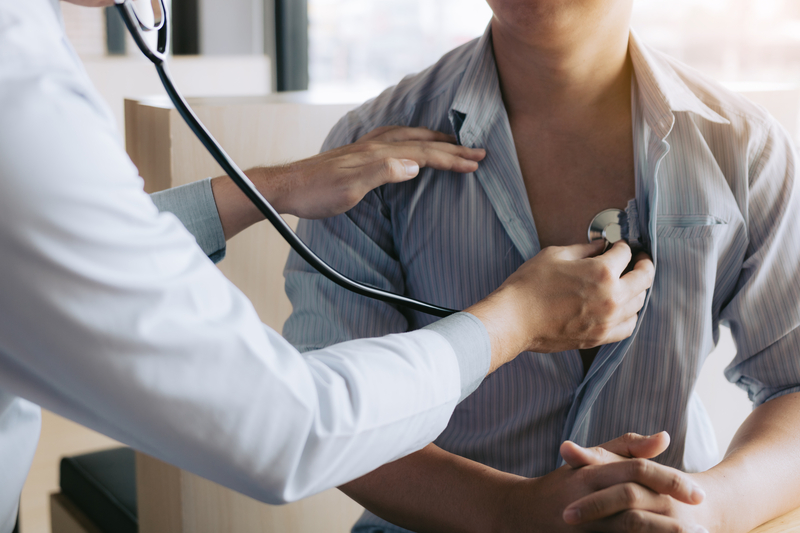 Health care worker's arm holding stethoscope against patient's chest, unbuttoned shirt.