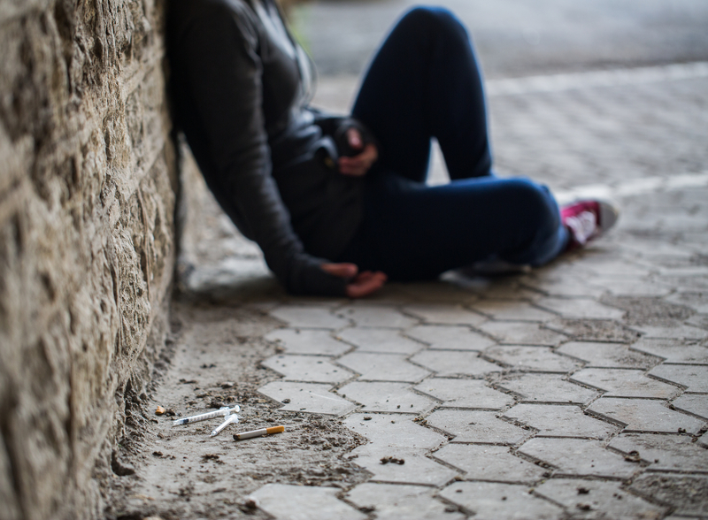 Woman sits against the wall, you can't see her head, drug paraphernalia near her.